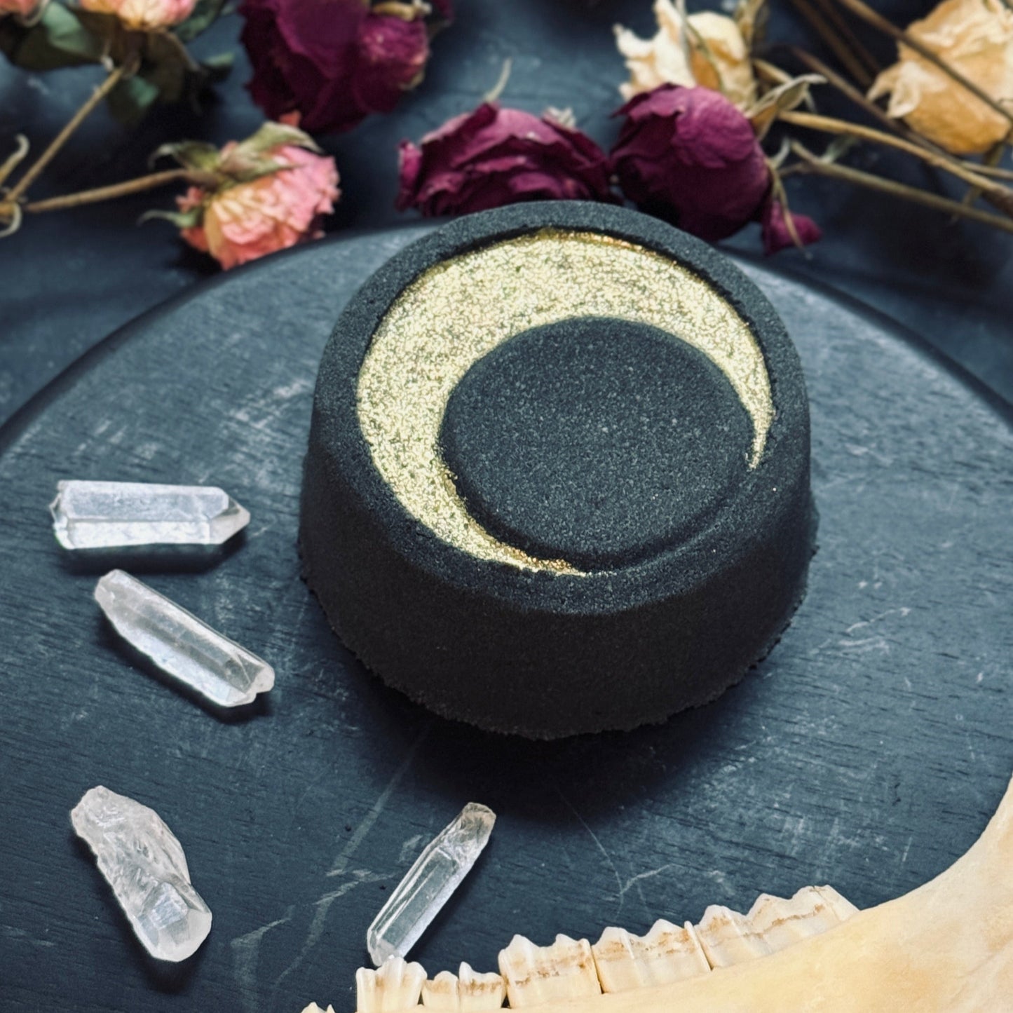 Round black bath bomb with gold crescent moon on a dark surface with crystals and dried flowers.