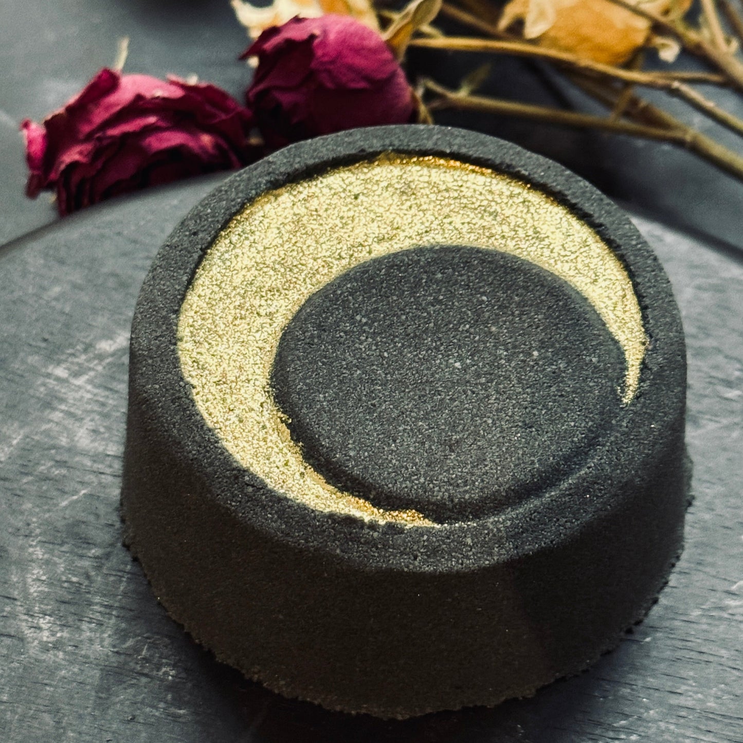 Round black bath bomb with a gold crescent moon with dried roses in the background.