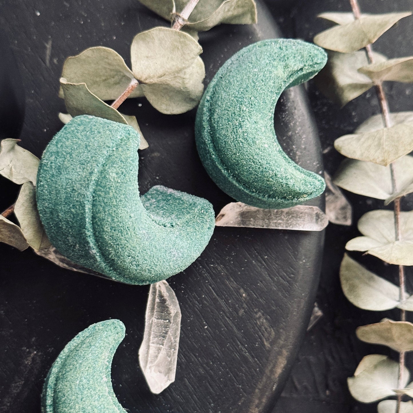 Image of crescent moon shaped green shower steamers with quart crystal points and dried eucalyptus leaves on dark background.