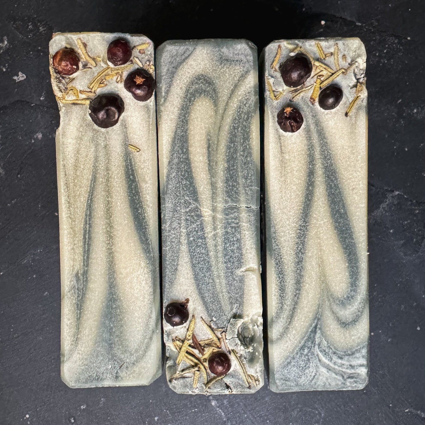 A top down view of 3 soap bars. Swirls of green and white with dried rosemary and juniper berries sprinkled along one side