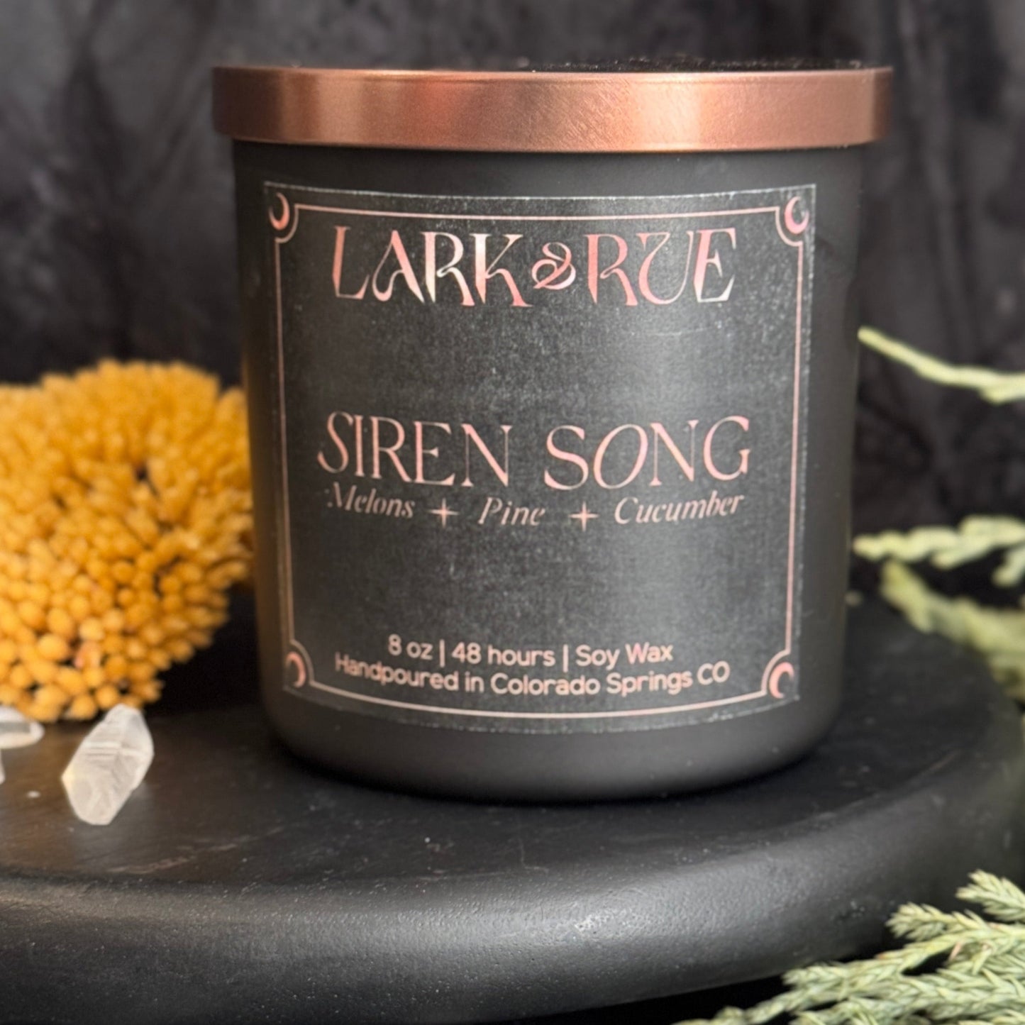 Siren Song on a dark surface with decorative evergreen foliage and clear quartz points.