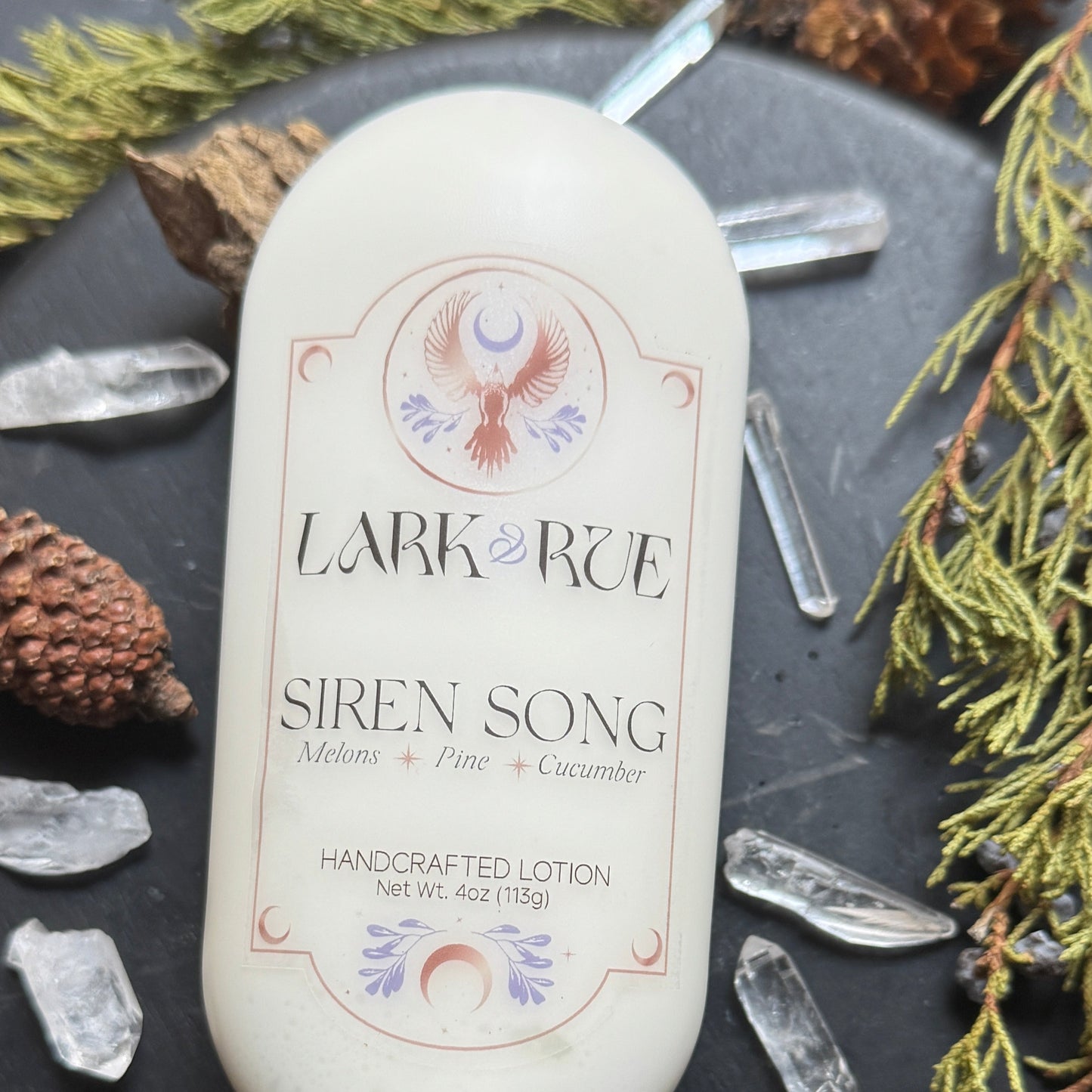 Siren Song lotion zoomed in on the bottle on a dark background surrounded by pine cones, evergreens, and quartz crystal points.