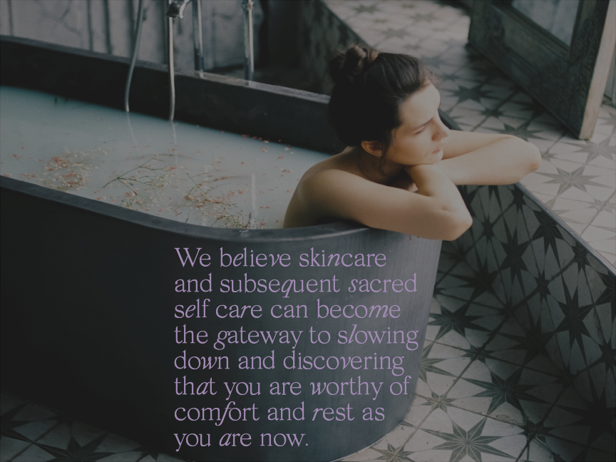 Image of a woman in a dark gray bathtub filled with creamy water with flowers on top. She is looking out the window resting her arms on the edge of the tub.