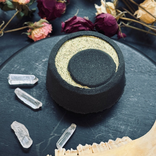 Round black bath bomb with gold crescent moon on a dark surface with crystals and dried flowers.