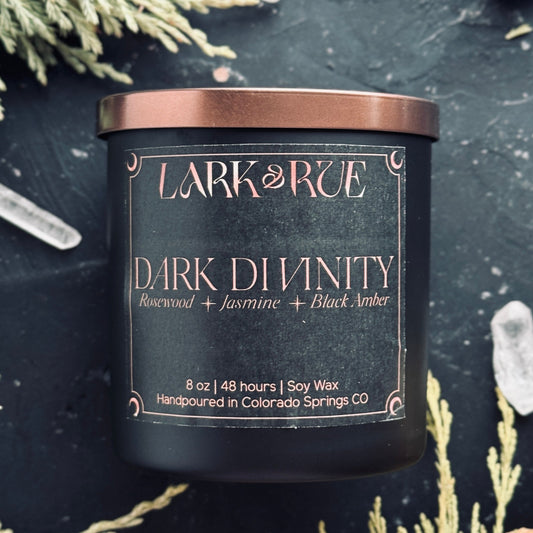 Dark Divinity candle on dark background with cedar sprigs and clear quartz points.