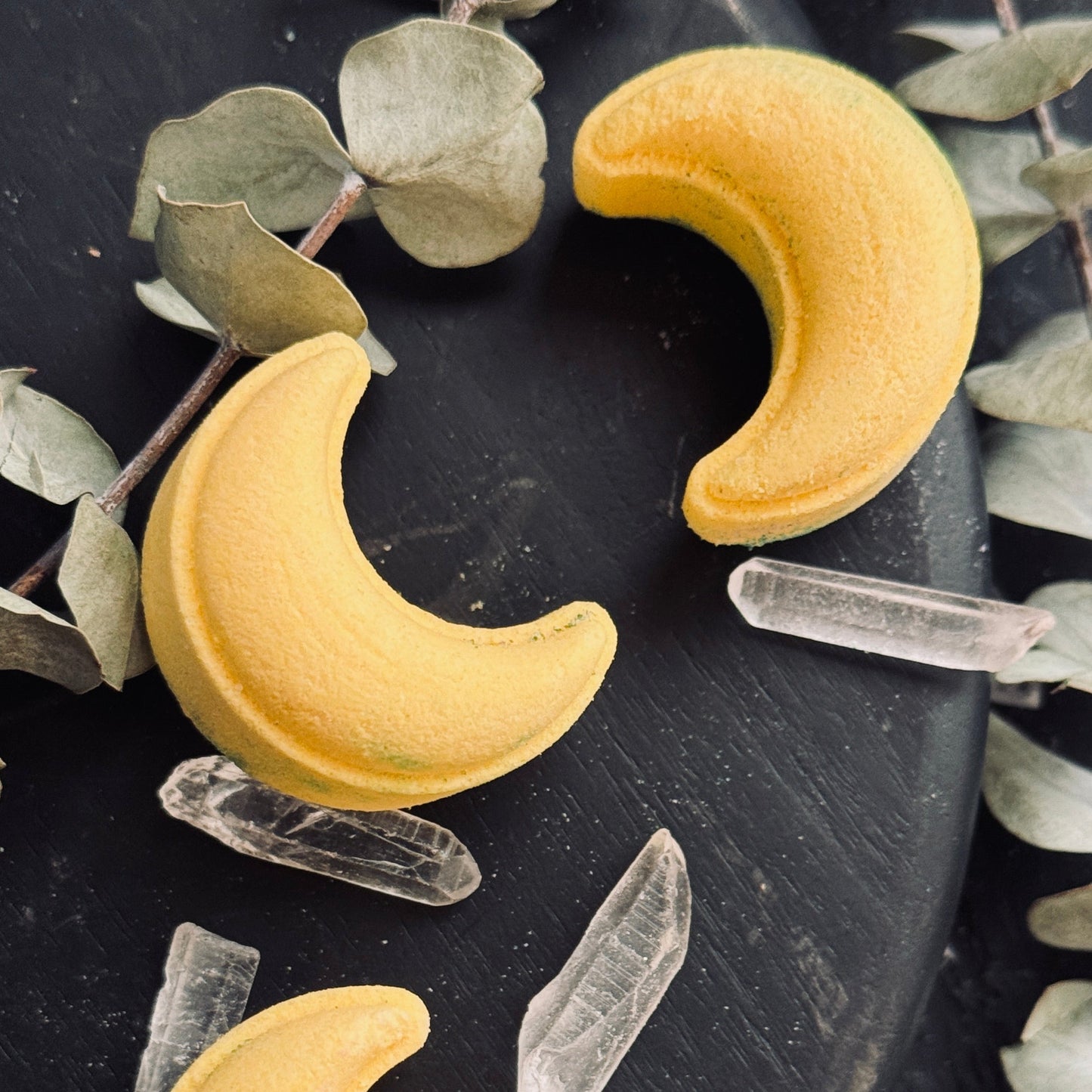 Image of yellow crescent moon shower steamers on dark background with quartz crystal points and dried eucalyptus leaves.