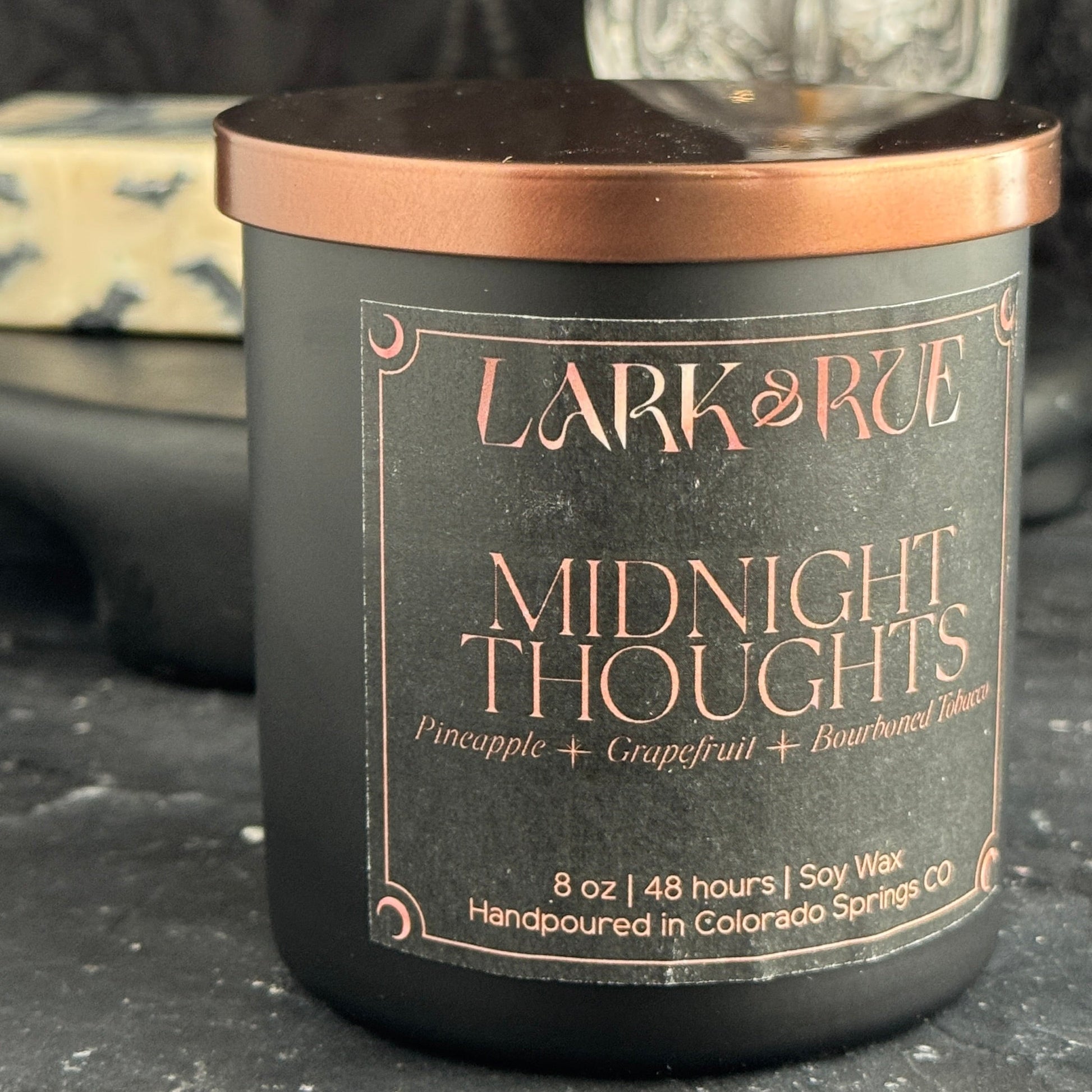 Midnight Thoughts candle on a dark surface with Midnight Thoughts soap in the background.