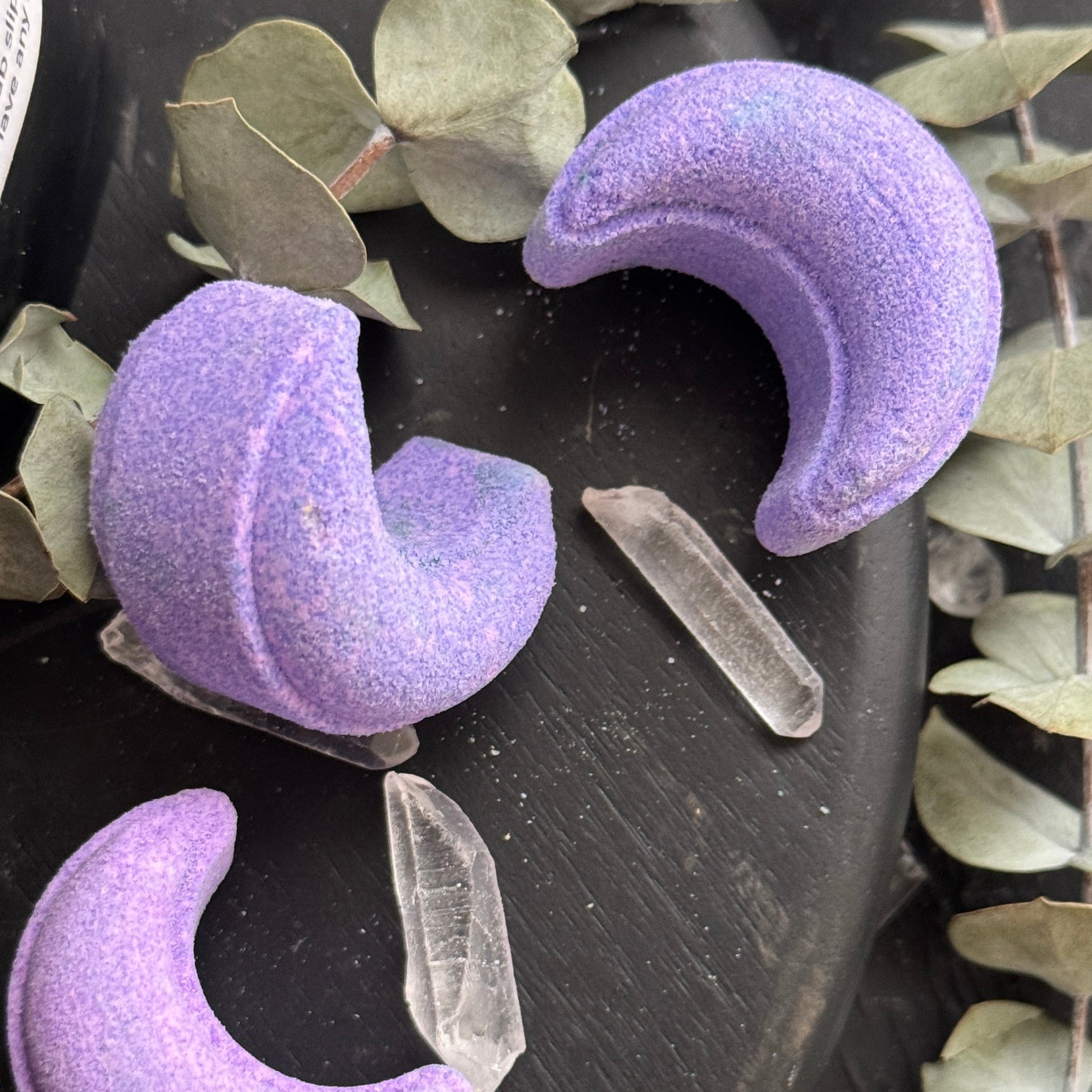 Image of light purple crescent moon shower steamers on dark background with quartz crystal points and dried eucalyptus leaves.