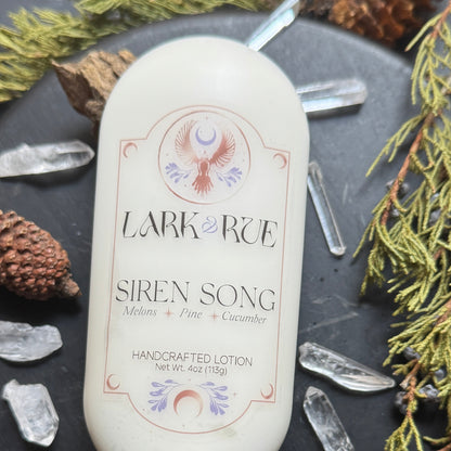Siren Song lotion zoomed in on the bottle on a dark background surrounded by pine cones, evergreens, and quartz crystal points.