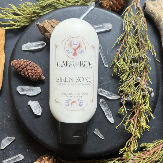 Siren Song Lotion on a dark background surrounded by pine cones, evergreens, drift wood, and quartz crystal points.